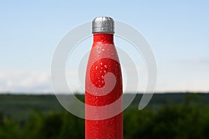 Thermo stainless bottle, sprayed with water. Sky and forest on background. Thermos of matte red color.