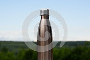 Thermo stainless bottle for water, tea or coffee. Sky and forest on background. On the glass desk. Thermos of maro color.
