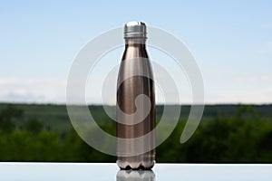 Thermo stainless bottle for water, tea or coffee. Sky and forest on background. On the glass desk. Thermos of maro color.