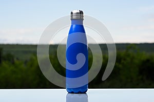 Thermo stainless bottle for water, tea or coffee. Sky and forest on background. On the glass desk. Thermos of matte blue color.