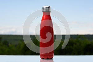 Thermo stainless bottle for water, tea or coffee. Sky and forest on background. On the glass desk. Thermos of matte red color.