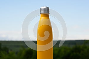 Thermo stainless bottle for water, tea or coffee. Sky and forest on background. On the glass desk. Thermos of matte yellow color.