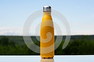 Thermo stainless bottle for water, tea or coffee. Sky and forest on background. On the glass desk. Thermos of matte yellow color.