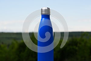 Thermo stainless bottle for water, tea or coffee. Sky and forest on background. Thermos of matte blue color.