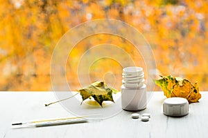 Thermometer, tablets from white bottle and yellow leaves on white table, with autumn colors on background. Cold and flu season