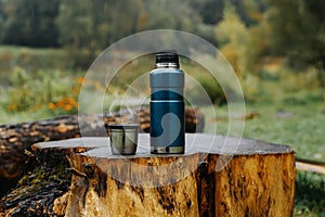 Thermos and aluminum hot drink mug with rising steam outdoors. Camping vacuum flask and iron cup standing on tree stump