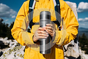 A thermos in the hands of a traveler. A man in tourist clothes with a thermos of coffee or tea in his hand against the background
