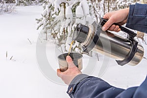 Thermos with hot tea or coffee in human hands against winter snow covered forest. Man pours drink into mug in cloudy winter day.