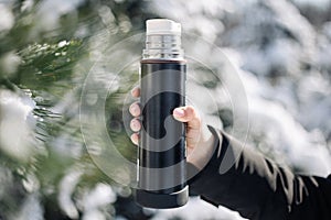 Thermos in the woman`s hand on a frosty cold winter day among snowed fir trees in the park. Closeup outdoors shot of a thermos