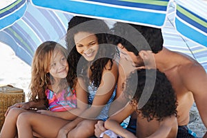 Theyre a close-knit family. A family of four sitting under their umbrella at the beach.