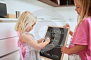 Theyre always willing to help with housework. two little girls writing a list of chores on a chalkboard at home.