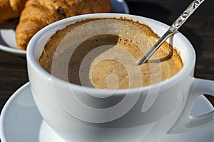 Thick foam of hot coffee cappuccino with spoon in white cup and croissant on wooden table, closeup. Milk foam cappuccino, latte