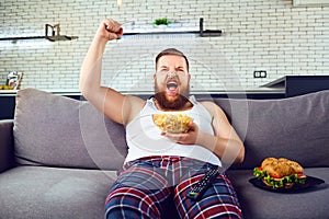 Thick funny man in pajamas eating a burger sitting on the couch.