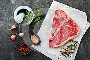 Thick Raw T-Bone Steak. Dry-aged Raw T-bone or porterhouse beef meat Steak on parchment paperwith herbs and salt on dark backgro