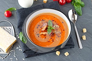 Thick tomato soup with basil and fried bacon in a black ceramic bowl on a grey abstract background. Healthy eating