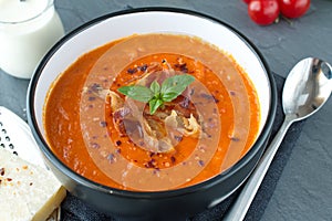 Thick tomato soup with basil and fried bacon in a black ceramic bowl on a grey abstract background. Healthy eating