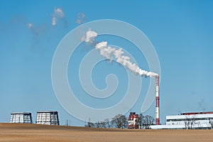 Thick white smoke coming out of a large pipe of an industrial facility or factory. A smoking chimney against a blue sky.