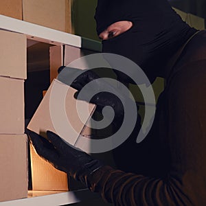 A thief in a black hood steals a parcel in a warehouse in the dark. Concept of problems with theft of goods and postal parcels