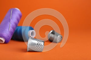 Thimbles, spools of sewing threads and needle on orange background, space for text