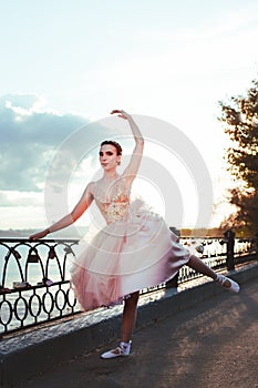 a thin ballerina in a pink silk corset dress gracefully raises her pointe-toed leg back on the river embankment in a photo