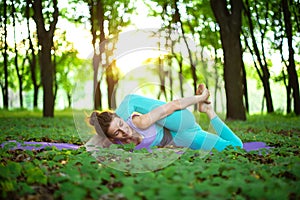 Thin brunette girl plays sports and performs yoga poses in a summer park on a sunset background. Woman doing exercises on the yoga