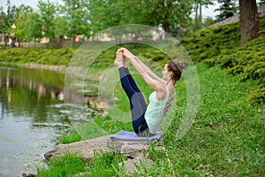 Thin brunette girl sitting in Both big toe exercise, Paripurna Navasana pose  in a summer park. Green forest on the background.