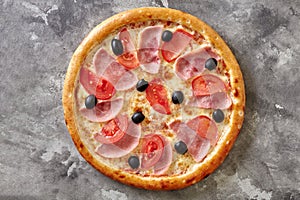 Thin classic pizza with melted cheese, ham, tomatoes and black olives on gray stone surface