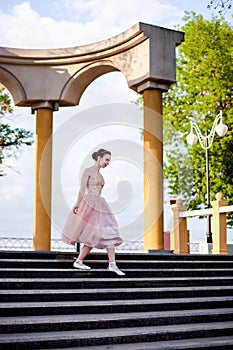 a thin female ballerina in a pink silk dress and pointe shoes with ribbons gracefully descends down the steps in an arch