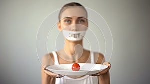 Thin girl with taped mouth holding plate with tomato, exhausting diet, anorexia photo