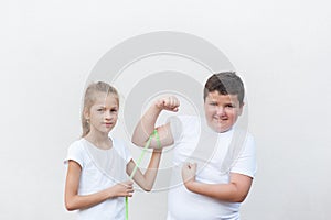 Thin little girl in white jersey measuring fat thick smiling boy muscle with tape on bright background