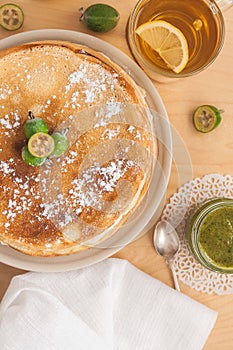 thin pancakes on a plate decorated with icing sugar, feijoa jam in a glass transparent small jar and a cup of green tea