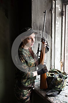 A thin red-haired girl stands at the broken window with a rifle. The girl in green camouflage with a gun. Military service for