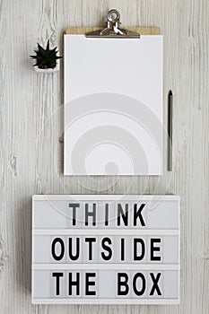 `Think outside the box` words on a light box, clipboard with blank sheet of paper over white wooden background, top view. Flat l