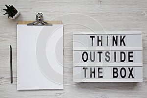 `Think outside the box` words on a light box, clipboard with blank sheet of paper over white wooden surface, top view. Flat lay,