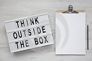 `Think outside the box` words on a light box, clipboard with blank sheet of paper on a white wooden background, top view. Flat l