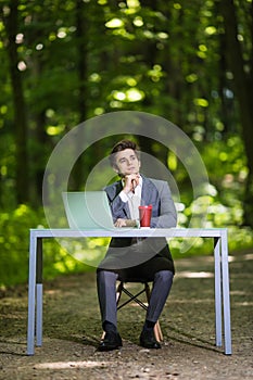 Thinking Businessman with hands on chin sitting at the office desk work at laptop computer and cup of coffee on road of green fore