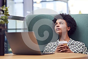 Thinking Businesswoman Using Laptop Working At Table In Breakout Seating Area Of Office Building 