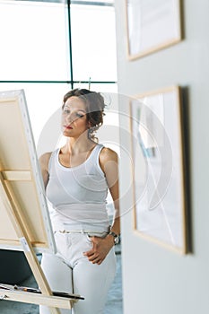 Thinking charming young woman artist in casual clothes with paint brushes in front of easel in modern art gallery