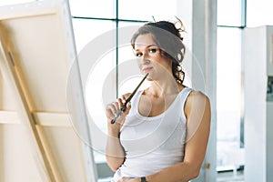 Thinking charming young woman artist in casual clothes with paint brushes in front of easel in modern art gallery