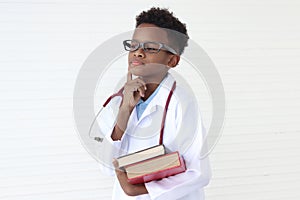 Thinking cute African boy in lab coat with stethoscope wearing glasses, holding pile of books on white wall room. Serious kid