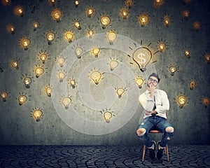 Thinking freelancer man sitting on chair looking up at many dollar idea light bulbs above head