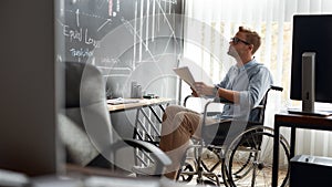 Thinking about future. Portrait of focused young male trader in a wheelchair holding some documents and looking at