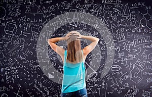 Thinking girl holding head against big blackboard, back view