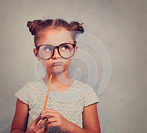 Thinking grimacing kid girl in glasses looking and holding pencil in hand on blue background with empty copy space. Toned vintage