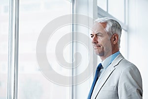 Thinking about his business future. A mature businessman standing and looking out of the window.