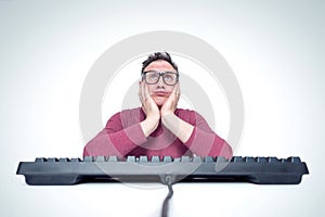 Thinking man in glasses behind a keyboard in front of a computer, holding his head in his hands. Dreaming programmer.