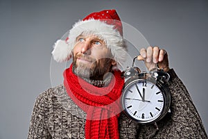 Thinking mature man wearing Santa hat holding big alarm clock looking up