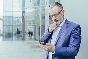 Thinking senior gray-haired investor reading news on phone, businessman standing outside office