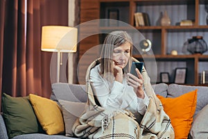 Thinking senior gray-haired woman at home sitting on sofa looking at phone screen