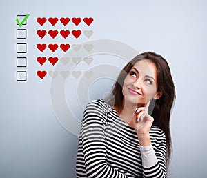 Thinking smiling young woman looking up and voting on five red hearts ranking on blue background